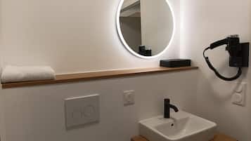 A modern bathroom with a white sink, a mirror with a light, a towel on a shelf, and a hairdryer on the wall.