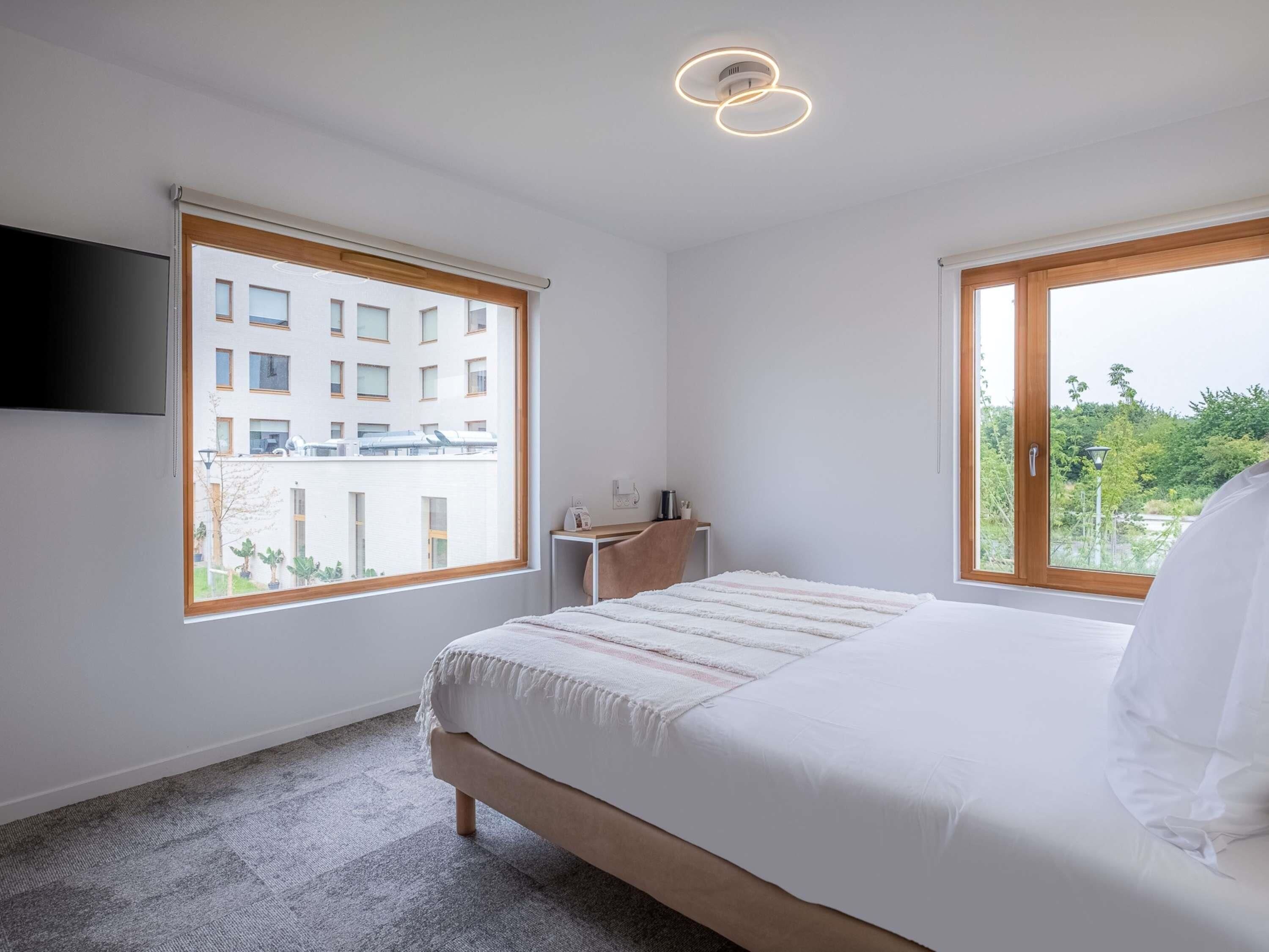 A bedroom with a bed, a bedside table, a TV mounted on the wall, and large windows with a view of a building and greenery.