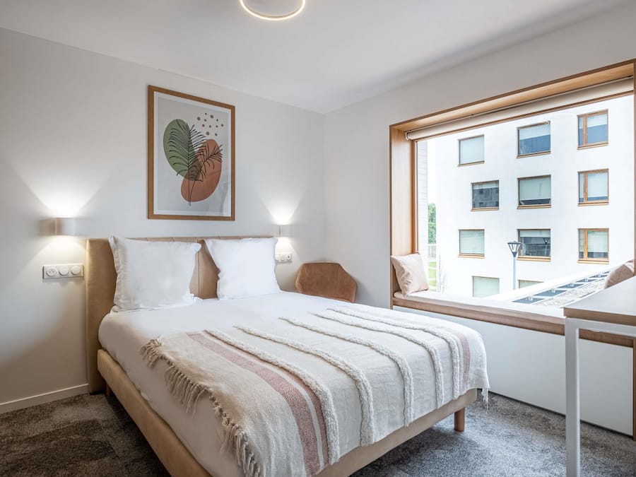 Une chambre à coucher avec un lit, des lampes de chevet, une fenêtre donnant sur un immeuble et une œuvre d’art encadrée accrochée au mur.