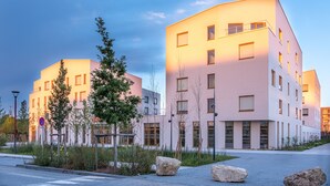 A modern building with multiple floors, large windows, and a flat roof. There are trees and rocks in the foreground. - ibis Styles Amiens Sud (Amiens)
