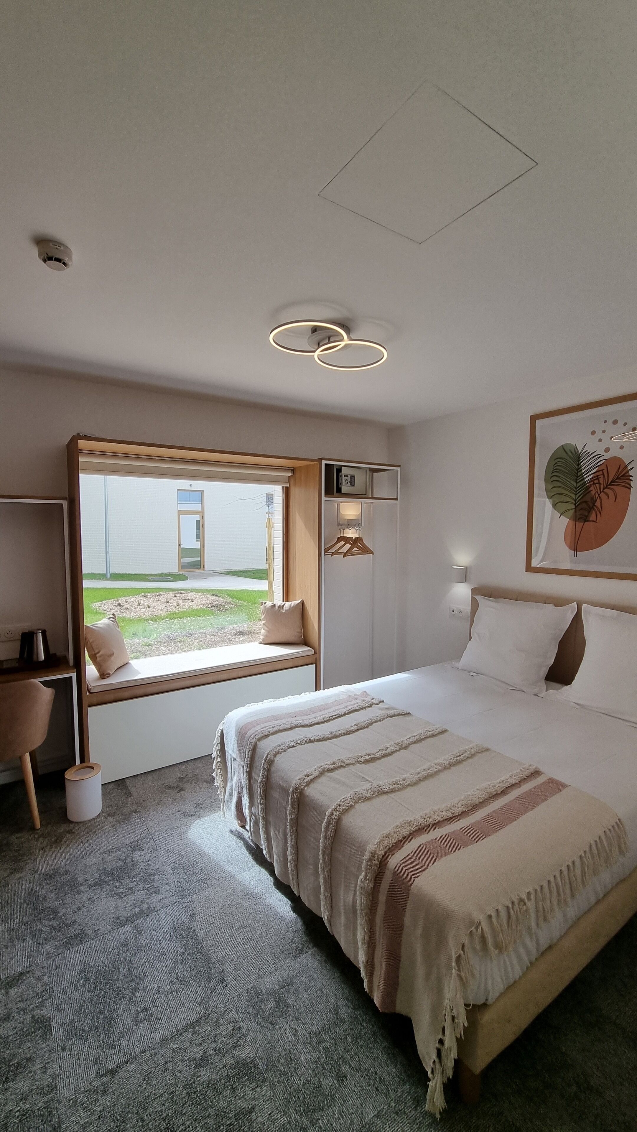 A modern bedroom with a large bed, a window seat, a wardrobe, and a ceiling light fixture.