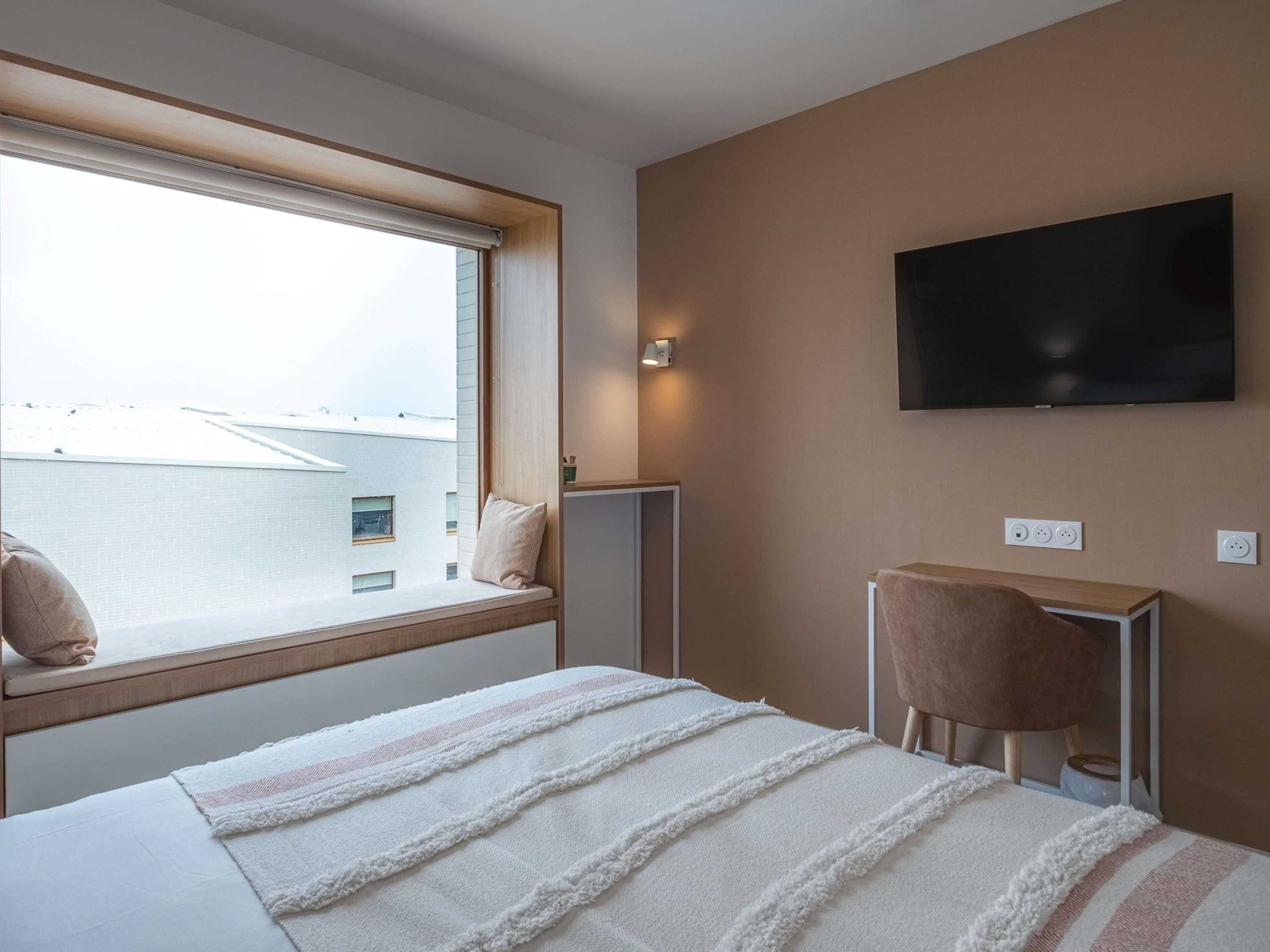 A hotel room with a bed, a TV mounted on the wall, a window with a view of buildings, and a small table with a chair.