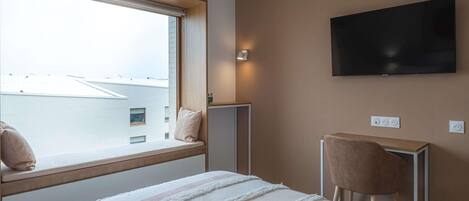 A hotel room with a bed, a TV mounted on the wall, a window with a view of buildings, and a small table with a chair.