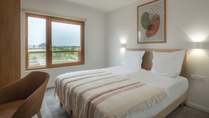 A bedroom with a large bed, a window with a view, a chair, and a wall-mounted light fixture. - ibis Styles Amiens Sud (Amiens)