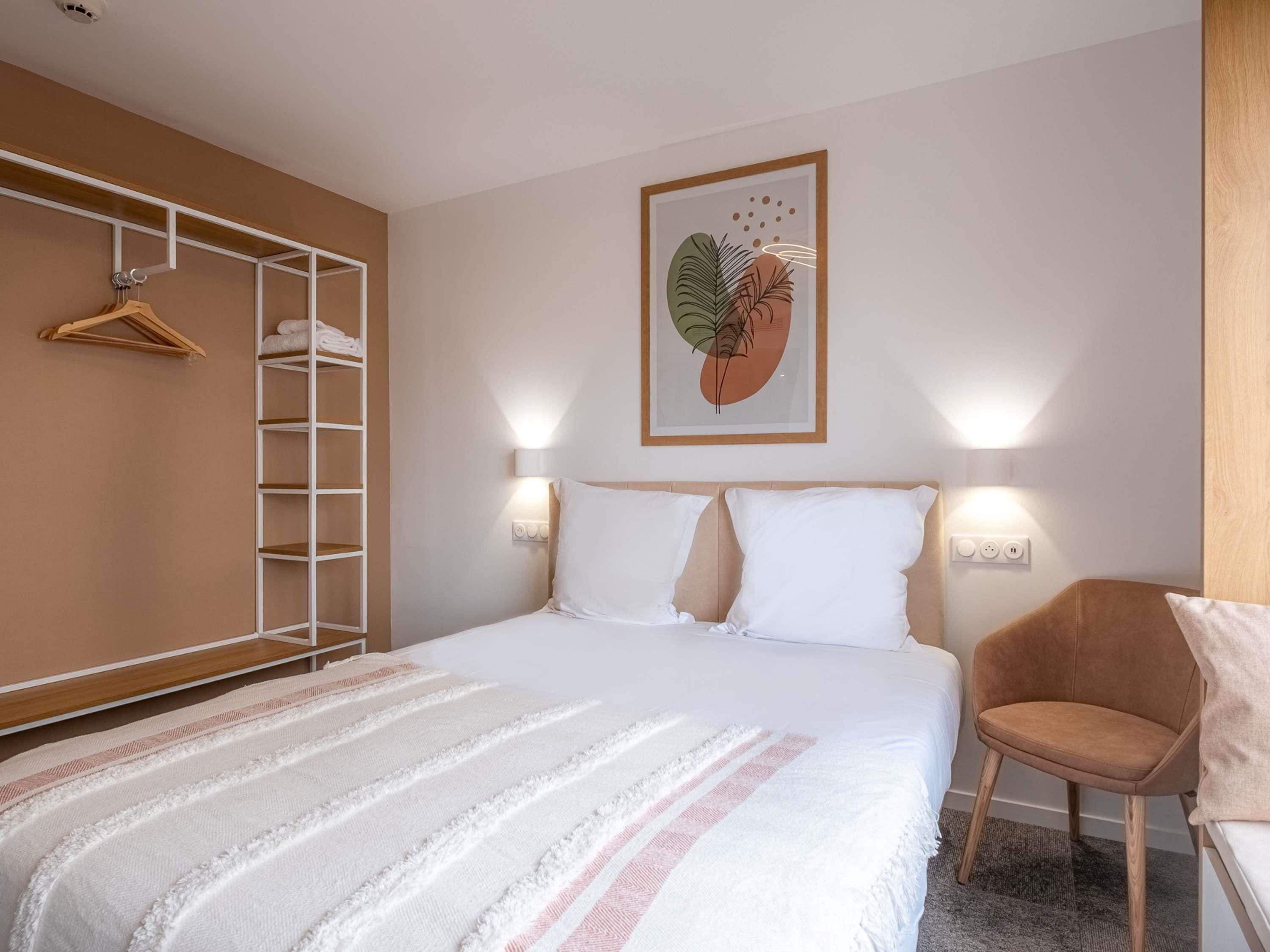 Une chambre d’hôtel comprenant un lit, une armoire, une chaise et un luminaire fixé au mur.