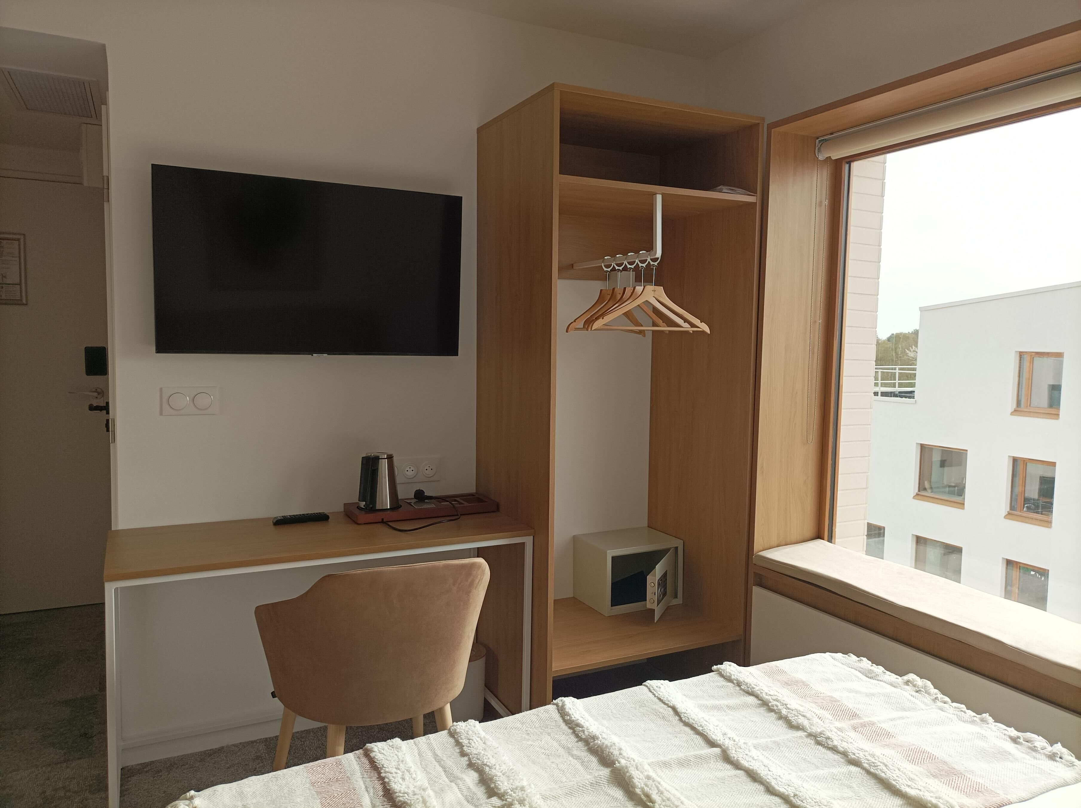 A bedroom with a bed, a desk with a chair, a TV, a wardrobe, and a window with a view of buildings.