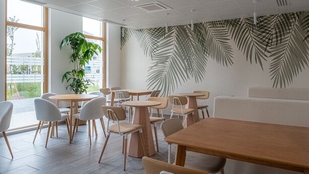 A modern cafe interior with wooden tables and chairs, large windows, and a wall decorated with palm leaf patterns. - ibis Styles Amiens Sud (Amiens)
