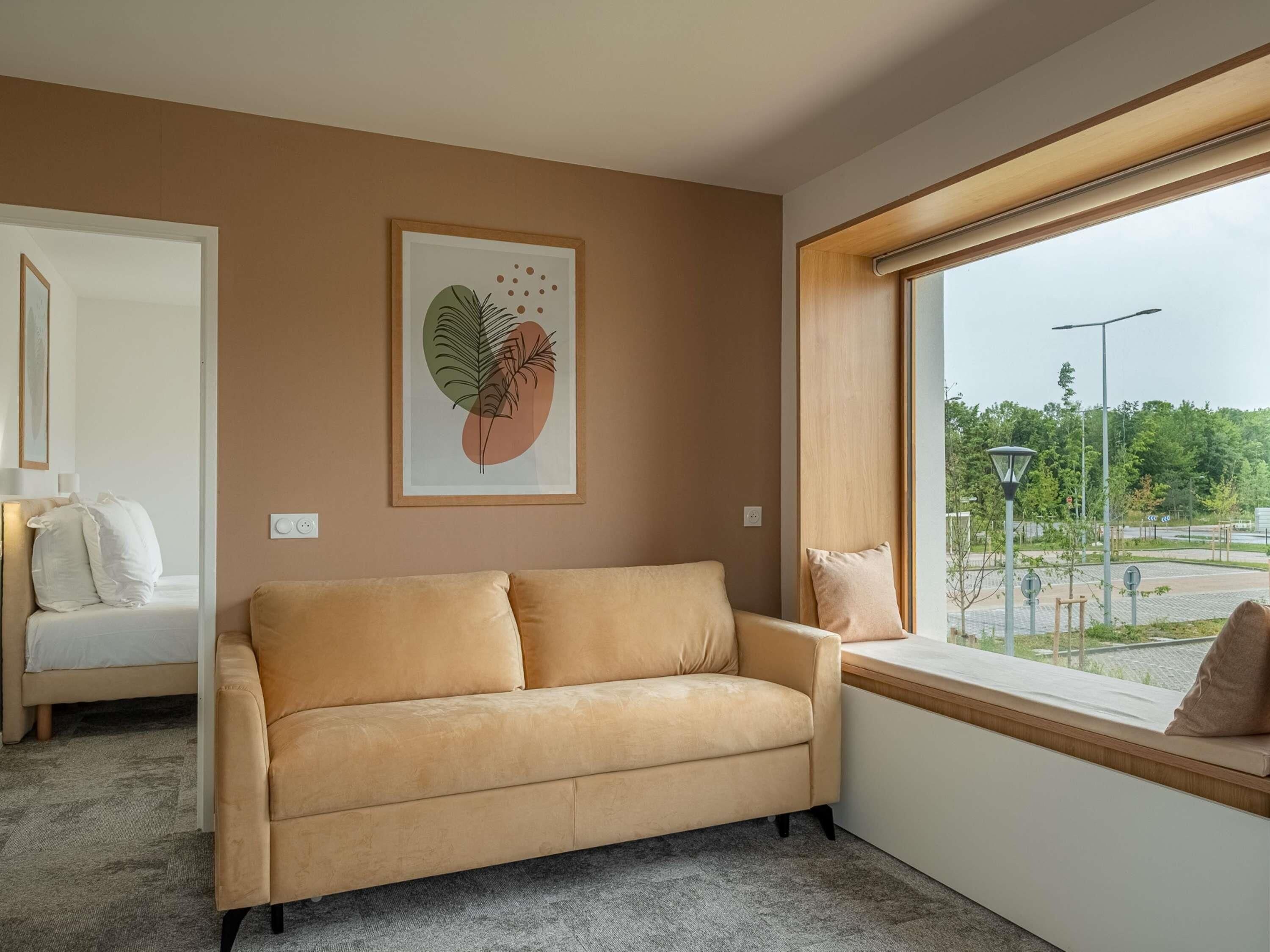 A modern living room with a beige sofa, a framed artwork on the wall, and a large window offering a view of a landscaped area.