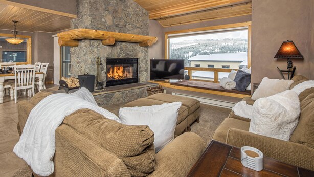 A cozy living room with a stone fireplace, comfortable sofas, a television, and a view of snowy mountains.