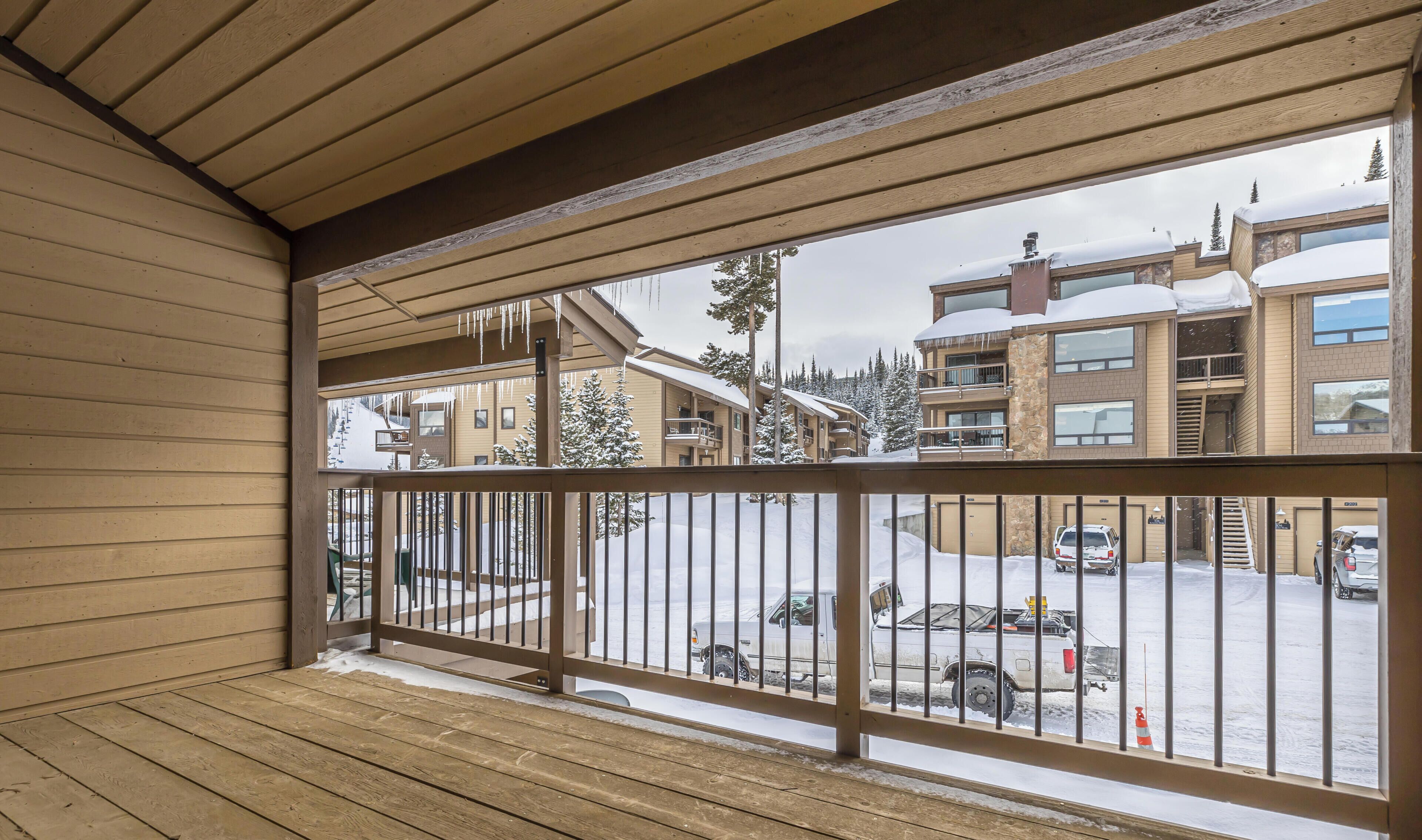 A wooden balcony with a metal railing overlooking a parking area and buildings with snow-covered roofs.