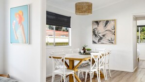 A dining area with a wooden table and white chairs, a large window with a view of a building, and two framed artworks on the walls. - Golden Sands - Pet Friendly - 2 Mins Walk to Beach (Culburra Beach)