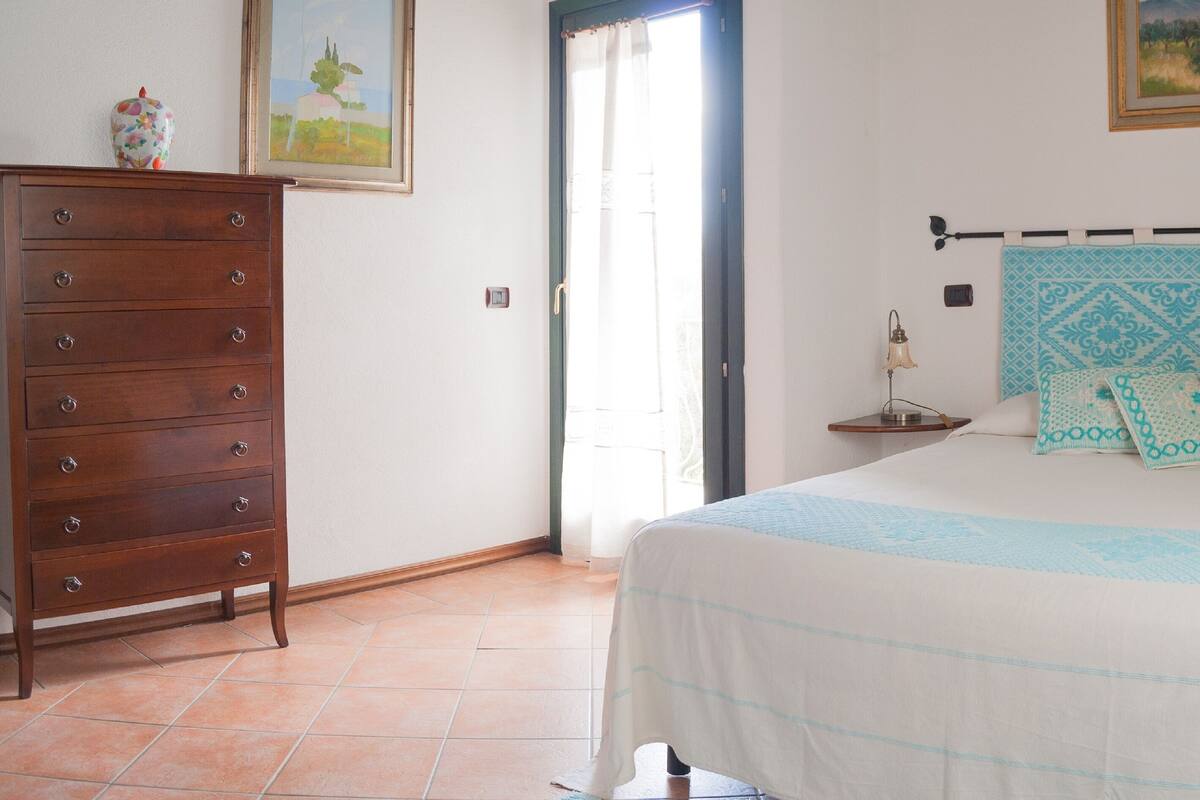A bedroom with a bed, a dresser, a nightstand, a lamp, and a window with curtains.