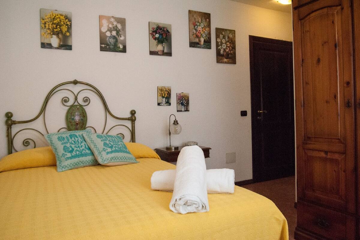 A neatly made bed with a yellow bedspread and teal pillows, a wooden wardrobe, and floral artwork on the wall.