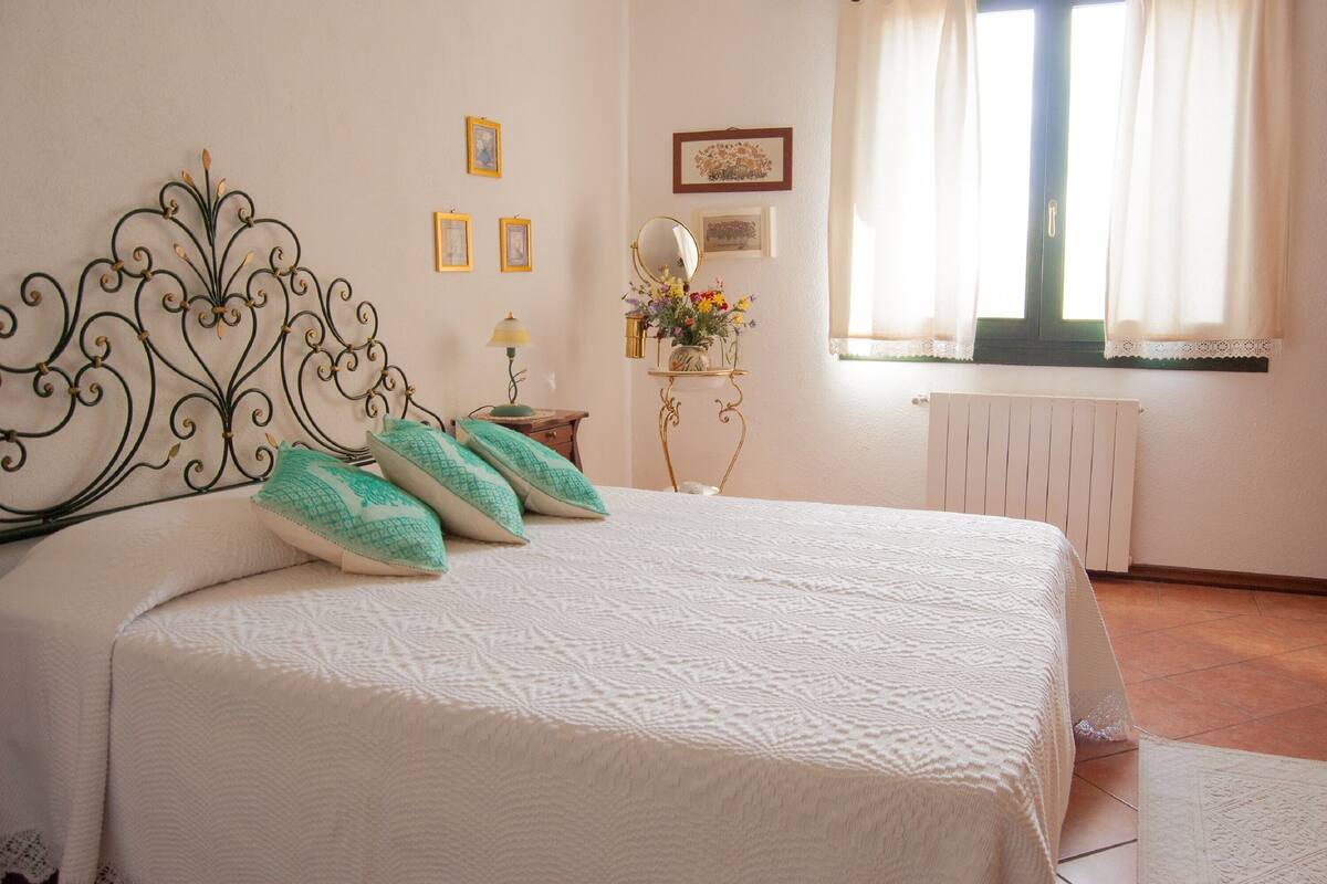 A neatly made bed with a decorative headboard, a bedside table with a lamp, and a window with curtains.
