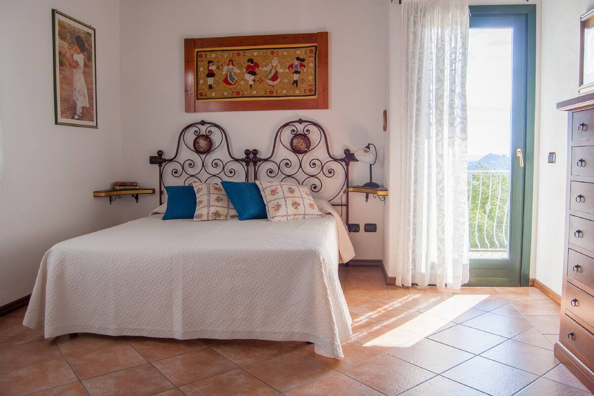 A bedroom with a bed, a nightstand, a dresser, a window with curtains, and a painting on the wall.