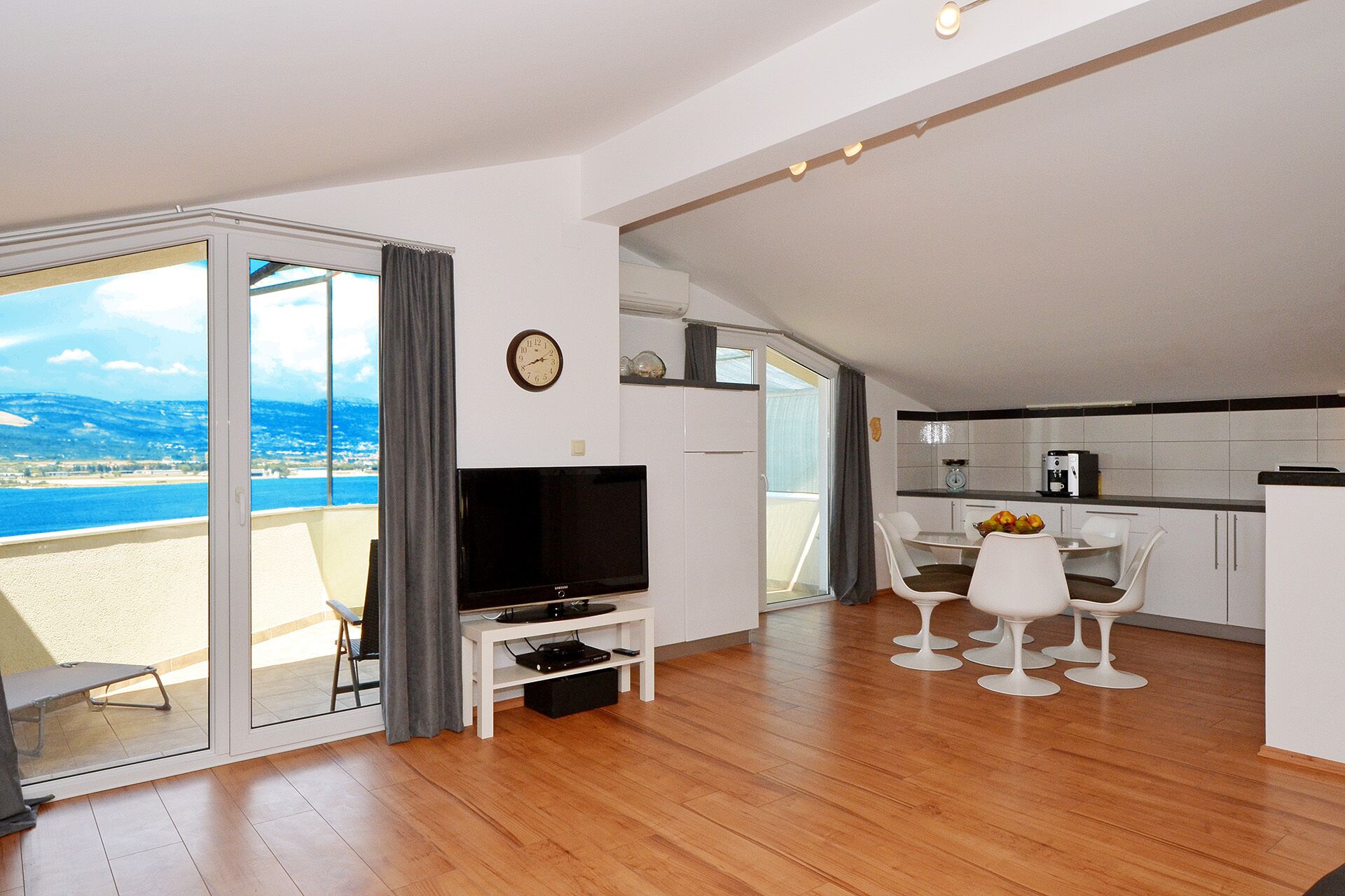 A modern living room with a television, a dining area with white chairs, and a kitchen with a coffee maker.