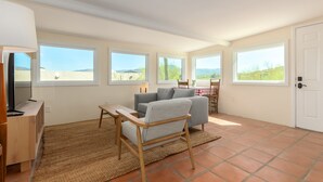 Un salon spacieux avec un canapé, une petite table et un téléviseur. De grandes fenêtres offrent une vue sur l’extérieur.