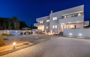 Ett modernt hus i flera våningar med en swimmingpool, utomhusmöbler och en väl upplyst entré.