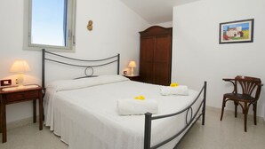 Un lit bien fait, avec du linge de lit blanc et deux serviettes, une chaise en bois, une table de chevet avec une lampe, une armoire en bois et un tableau encadré au mur.