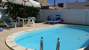 Une piscine dont le bord est recouvert de carreaux, deux échelles en métal, des chaises en plastique blanc, une table avec un parasol blanc et une plante en pot.