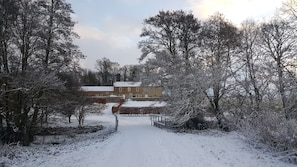 Ein verschneiter Pfad führt zu einem Ziegelgebäude mit schneebedecktem Dach, umgeben von kahl stehenden Bäumen.