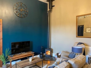 Uma sala de estar com parede azul escura, mesa de centro de madeira, sofá, televisão, lareira e um relógio.