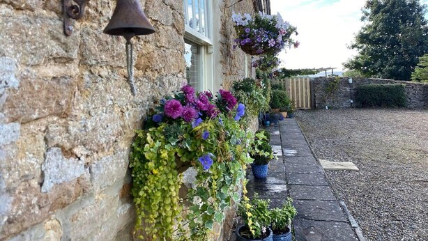 A stone building with a bell and a decorative metal piece. There are potted plants and flowers along the wall. - Luxury, Self-contained Country Cottage With View (Scarborough)