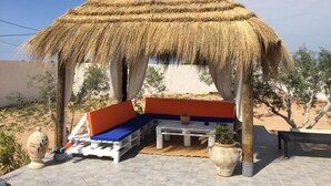 A thatched-roof structure with a bench and cushions, surrounded by potted plants and a clear sky.