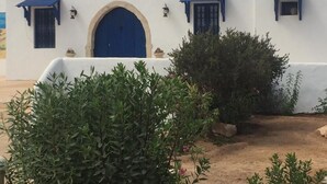 A white building with a blue door and windows, surrounded by greenery and a sandy ground.