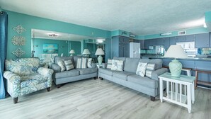 Una sala de estar con un sofá seccional gris, un sillón estampado y una mesita blanca con una lámpara.