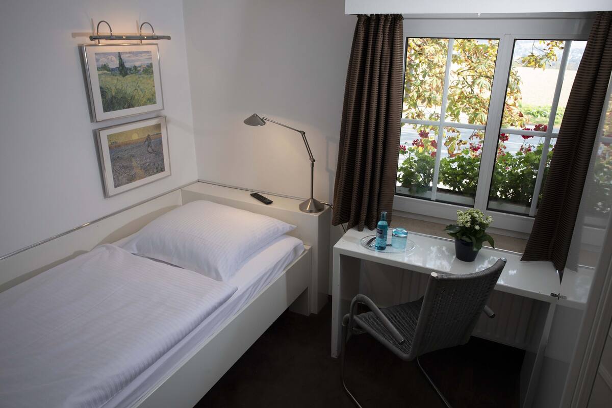A single bed with white linens, a desk with a chair, a lamp, and a window with curtains.