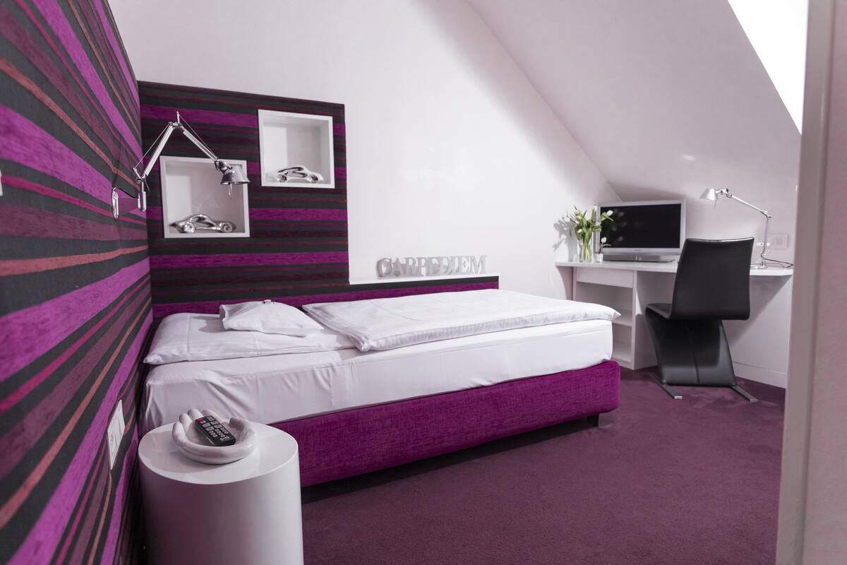 A modern bedroom with a sloped ceiling, a bed with white and purple bedding, a desk with a computer, and a chair.