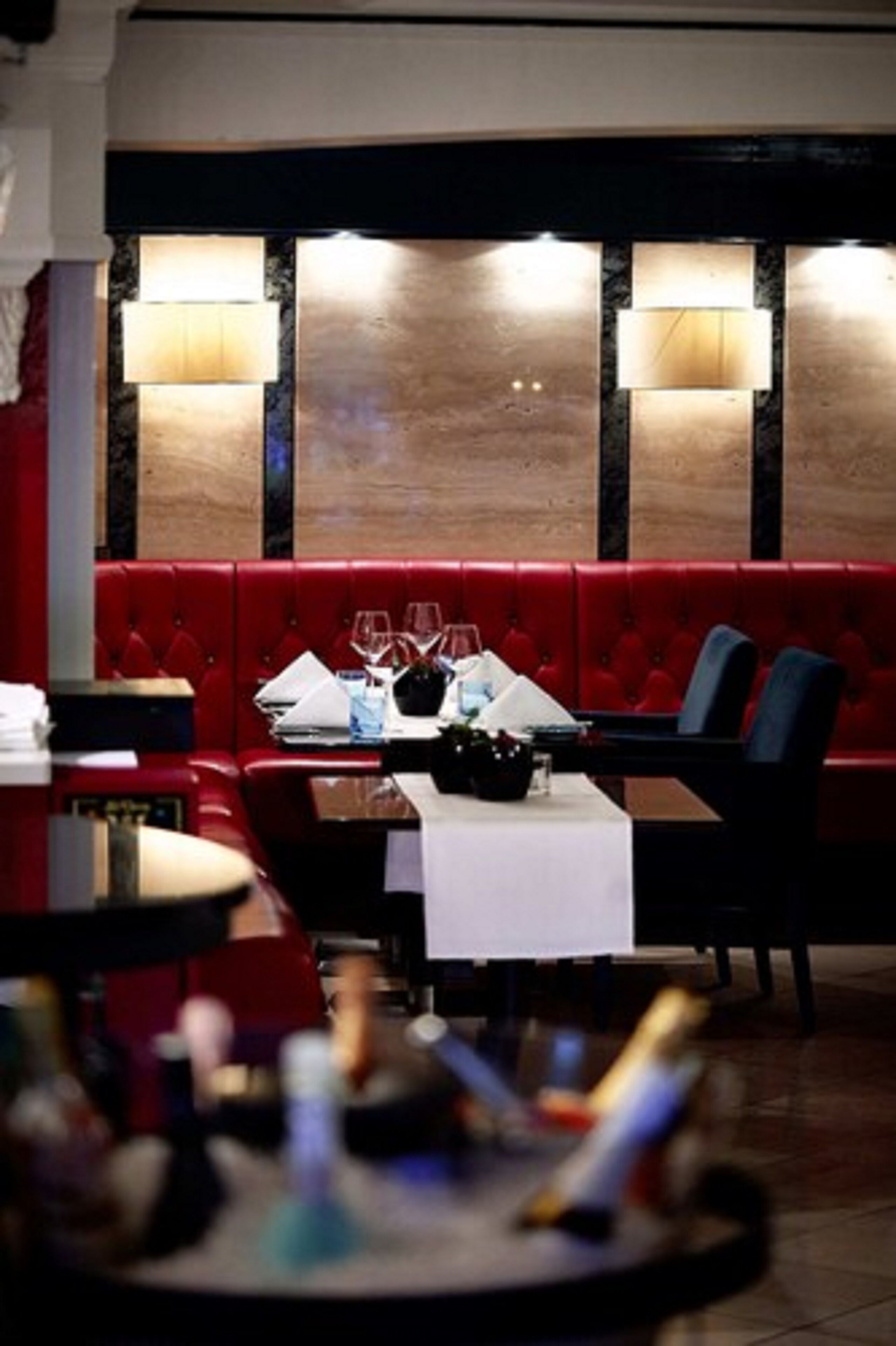 A restaurant interior with red upholstered booths, a table set with wine glasses and a bottle, and wall-mounted lamps.