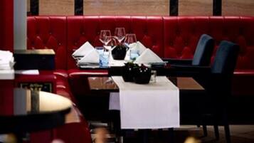 A restaurant interior with red upholstered booths, a table set with wine glasses and a bottle, and wall-mounted lamps.