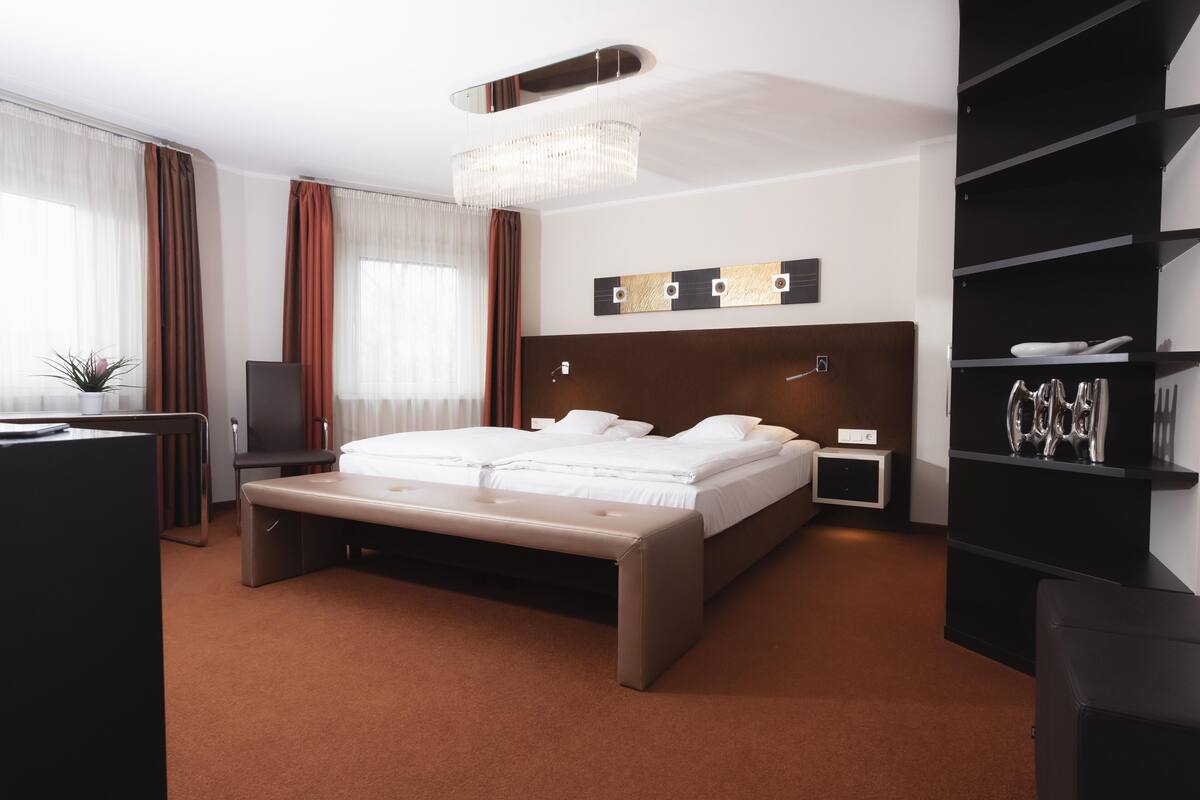A modern hotel room with a large bed, a bench, a desk, a bookshelf, and a chandelier.