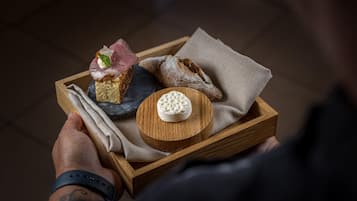 Una bandeja de madera con un trozo de pan, una rebanada de carne con guarnición y un queso redondo sobre una tabla de madera.