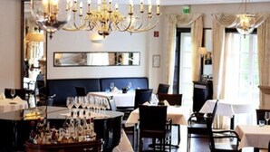 A dining area with a piano, tables, chairs, chandeliers, and a bar cart with bottles. - Haus Gerbens (Wickede)