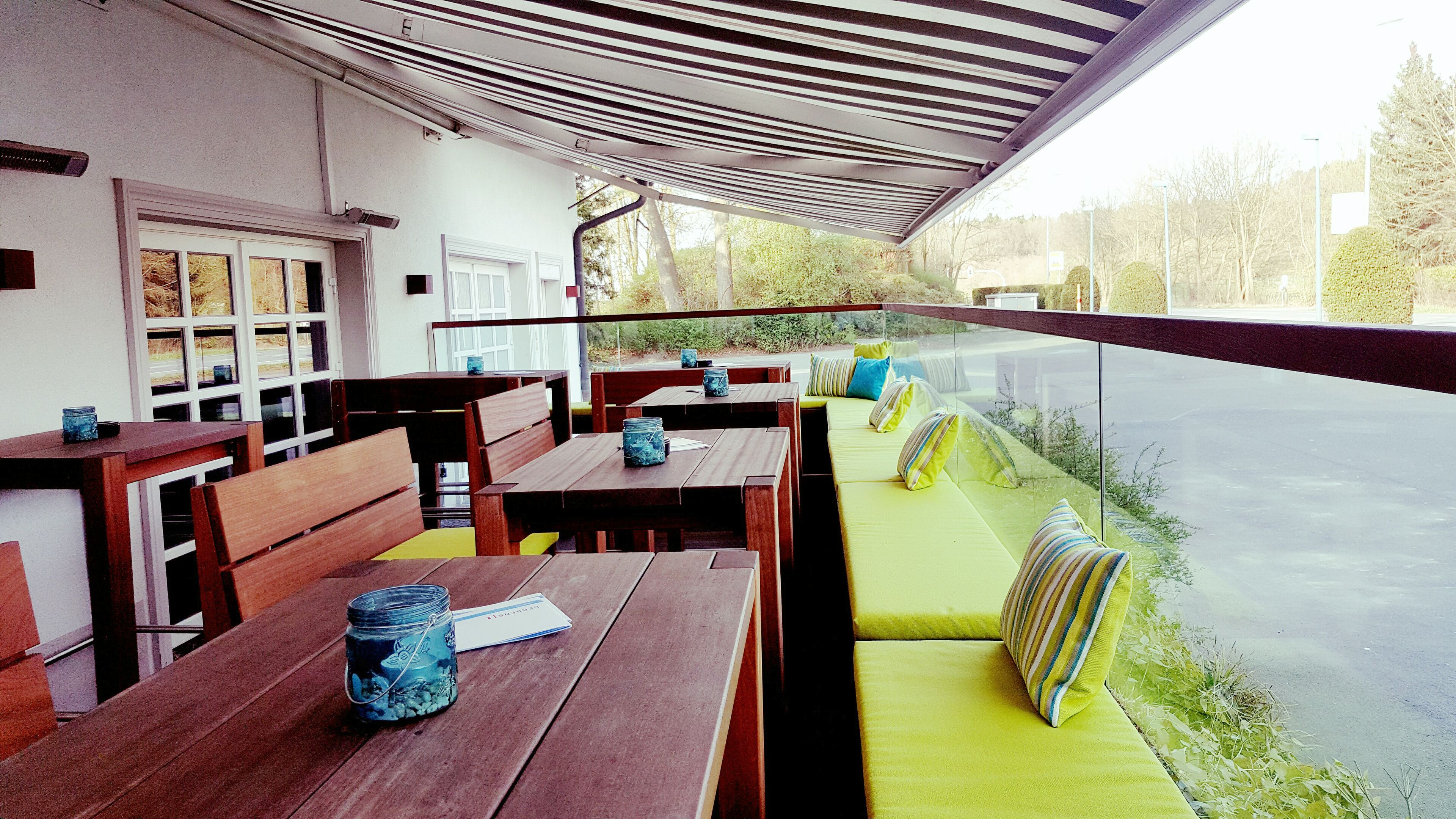 An outdoor seating area with wooden tables and benches, a green cushioned bench, and a view of a road and trees.