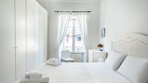 A hotel room with a neatly made bed, a chair, a small table with a vase, and a view of the outside through a window with curtains. - Bright apartment near Villa Borghese (Roma)