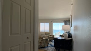 Een hotelkamer met een deur, een zithoek met twee stoelen, een tafel met een lamp en uitzicht op de oceaan door een raam.