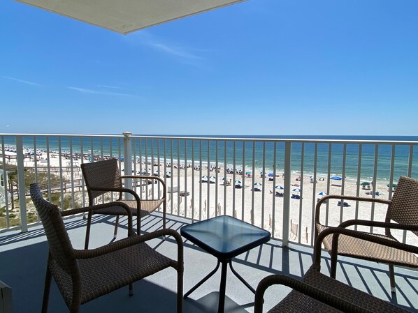 A balcony with wicker chairs and a glass table overlooking a beach with umbrellas and people. - Beachfront Gulf View Condo by PKRM (Perdido Key)