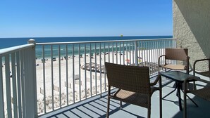 Een balkon met rieten stoelen en een tafel, met uitzicht op een strand met parasols en mensen.