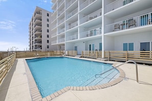 A swimming pool with a wooden deck and a white building with balconies in the background.