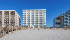 Een hotel aan het strand met meerdere balkons, een zandstrand ervoor en een helderblauwe lucht.