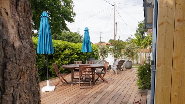Eine Holzterrasse mit Tisch und Stühlen, zwei blauen Sonnenschirmen und Kübelpflanzen.