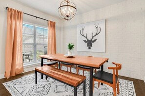 A dining area with a wooden table, four chairs, a bench, a chandelier, a wall-mounted deer head artwork, and a window with blinds.