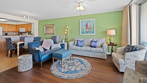 Ein Wohnzimmer mit blauem Sofa, einem gestreiften Sessel und einem Hocker, ein Essbereich mit Holztischen und -stühlen, eine Küche mit Holzausstattung und einem Deckenventilator.