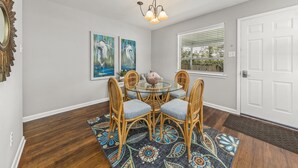 A dining area with a round glass table, four wicker chairs, two wall-mounted paintings, and a chandelier.