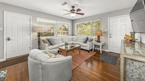 A living room with a sectional sofa, a coffee table, a ceiling fan, and a view of the outdoors through large windows.