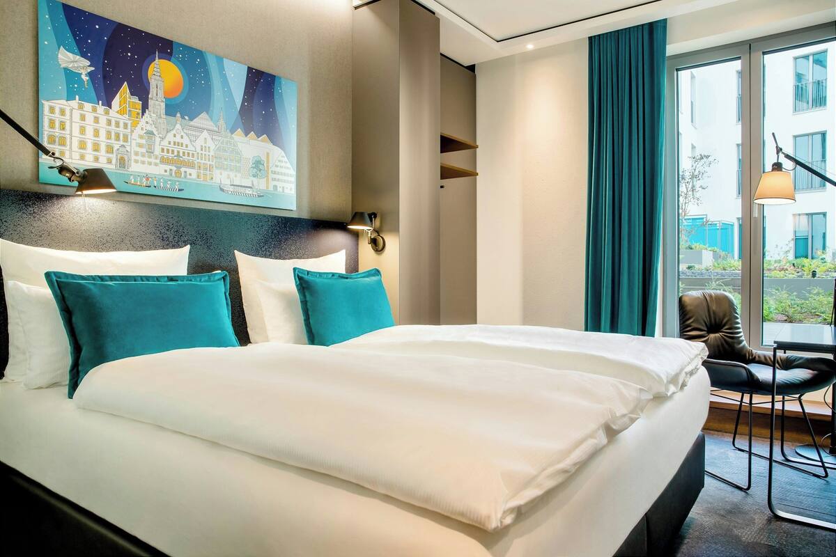A hotel room with a large bed, teal pillows, a cityscape painting, a view of the outdoors, and a chair by the window.